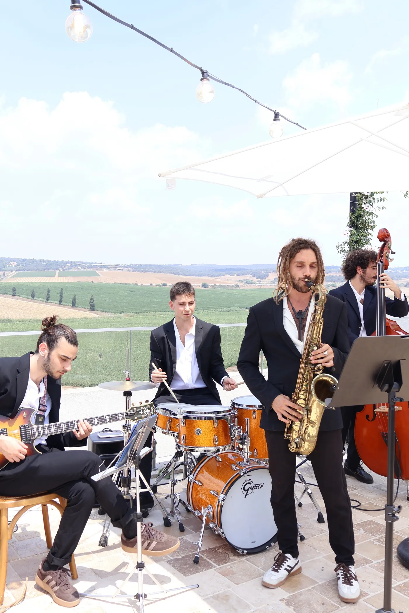 Cortado playing in daylight on a vineyard terrace — guitar, drums, saxophone and double bass under a white parasol, with green fields behind.