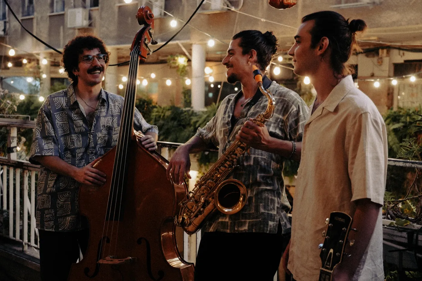 Three members of Cortado laughing together on a terrace at dusk beneath strings of warm lights, holding a double bass, a saxophone and a guitar.