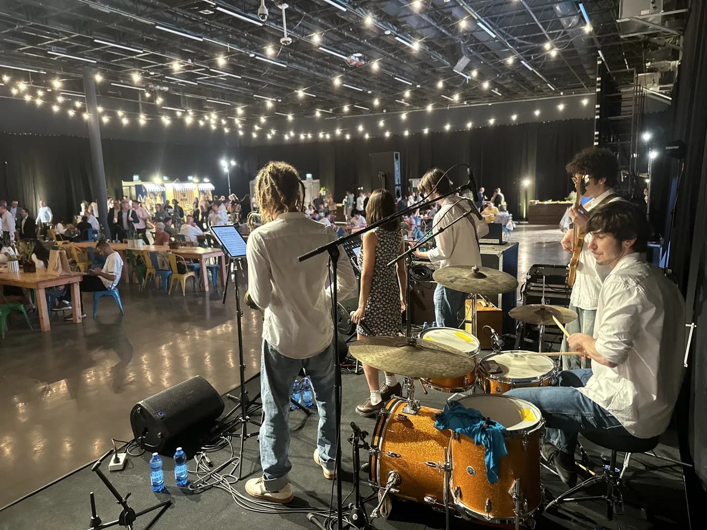 The band seen from the stage during a large indoor event, strings of lights overhead and several hundred guests seated beyond.